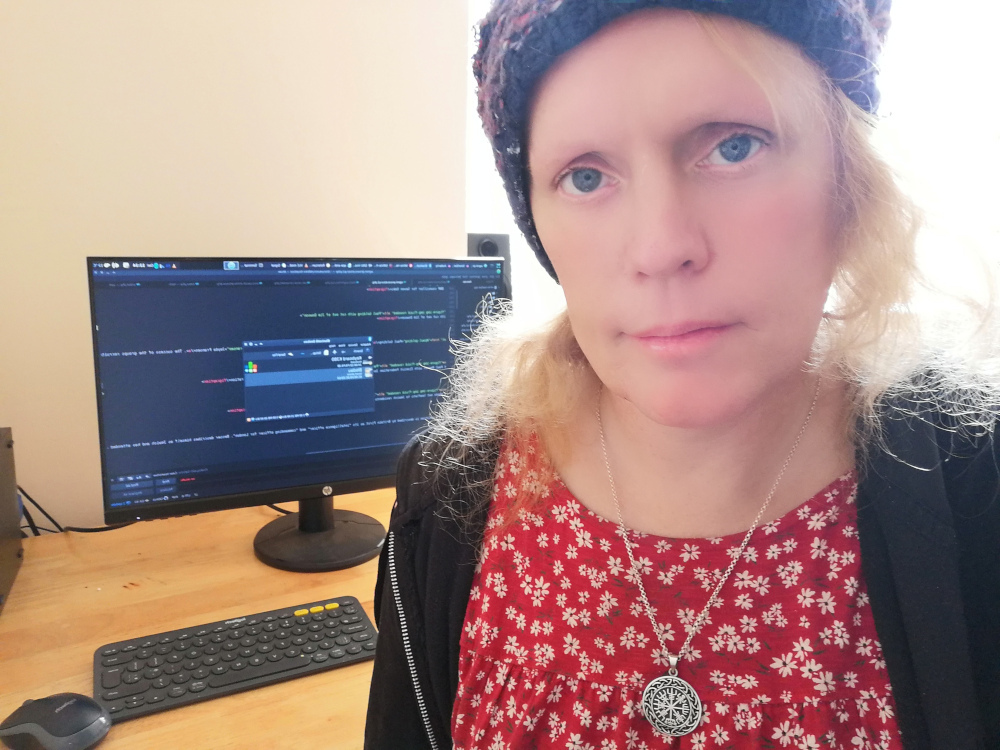 Léonie wearing a wolly red and blue hat, a red floral dress with a silver Vegvisir pendant seated leaning foward, behind a modern flat screen desktop computer 2020