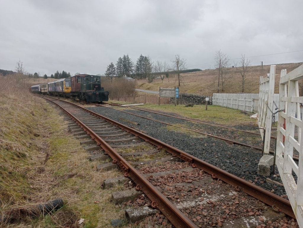 Steam engine, on disused tracks, behind a white roadside gate.